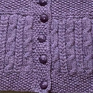 May include: Close-up of a lavender knitted cardigan with a textured cable knit design. The cardigan features a button-up front with matching purple buttons. The knitwear has a detailed pattern and a cozy, handmade aesthetic.