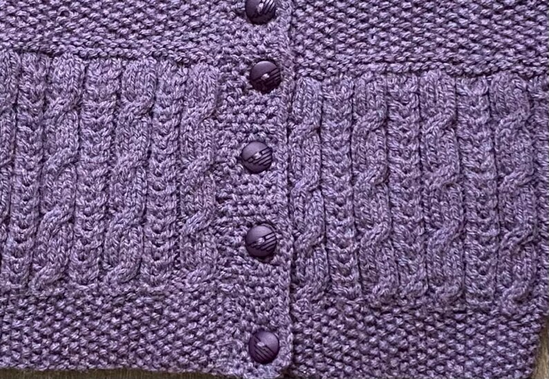 May include: Close-up of a lavender knitted cardigan with a textured cable knit design. The cardigan features a button-up front with matching purple buttons. The knitwear has a detailed pattern and a cozy, handmade aesthetic.