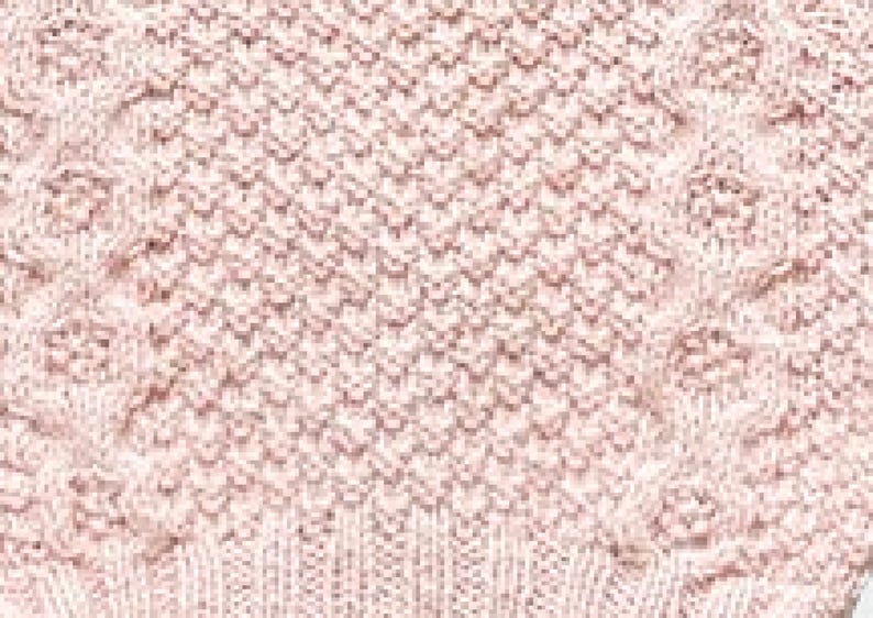 Op de afbeelding: Close-up van een gebreide stof in een zachte, gedempte roze tint. De textuur heeft een combinatie van kabel- en ruitpatronen, waardoor een visueel interessant oppervlak ontstaat. De onderrand toont een geribbeld breidetail.