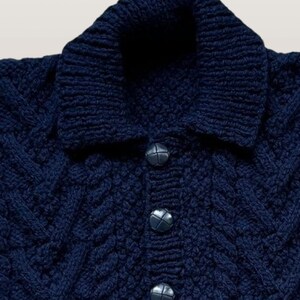 May include: A dark navy blue knit sweater featuring a thick folded collar, a vertical button placket with three round dark buttons, and a textured cable knit pattern throughout the fabric. The garment has a structured neckline and long sleeves.