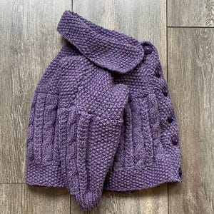 May include: A purple knitted cardigan with a large collar and cable knit detailing on the sleeves and front. The cardigan has a button-up closure with matching purple buttons. The garment is made of a soft, textured yarn.