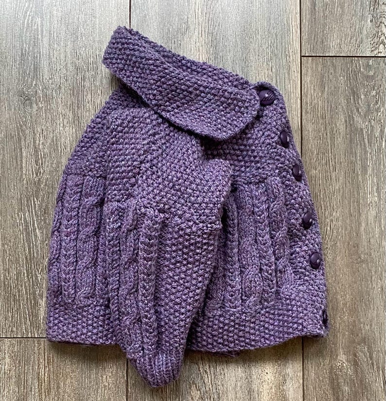 May include: A purple knitted cardigan with a large collar and cable knit detailing on the sleeves and front. The cardigan has a button-up closure with matching purple buttons. The garment is made of a soft, textured yarn.