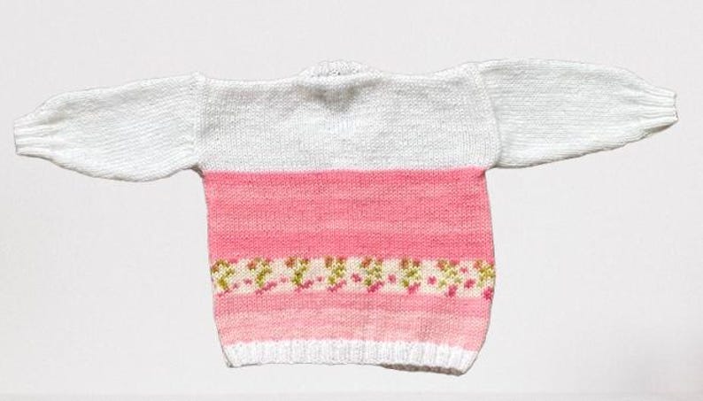 May include: A long-sleeved knit jumper featuring a white upper section and a pink lower section. The pink area includes horizontal stripes in varying shades of pink and a decorative band with a small floral pattern in green and pink hues.