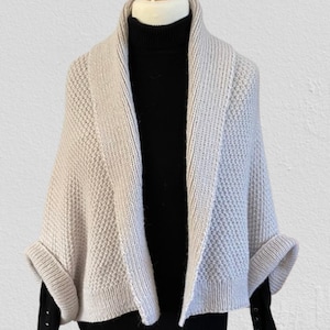 Op de afbeelding: Een beige gebreid vestje met een gestructureerd honingraatpatroon en een brede geribbelde sjaalkraag. Het kledingstuk heeft korte mouwen met omslag en wordt gedragen over een zwart shirt met lange mouwen en een col met zichtbare knopen.