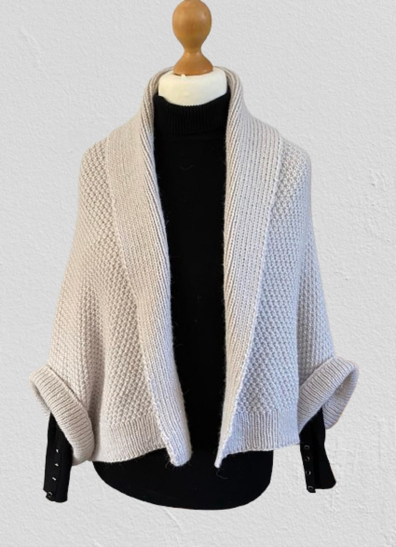 Op de afbeelding: Een beige gebreid vestje met een gestructureerd honingraatpatroon en een brede geribbelde sjaalkraag. Het kledingstuk heeft korte mouwen met omslag en wordt gedragen over een zwart shirt met lange mouwen en een col met zichtbare knopen.
