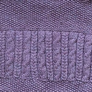 May include: Close-up of a knitted fabric in shades of purple. The top and bottom sections have a textured, tight knit, while the middle section features a cable knit design. The overall texture is soft and detailed.