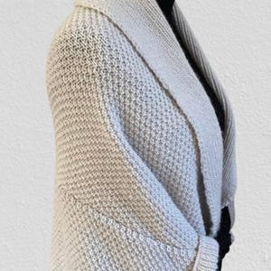 Op de afbeelding: Een beige gebreid vest met een brede sjaalkraag en een gestructureerd breipatroon. Het kledingstuk heeft lange mouwen met geribbelde boorden en een zoom die op de taille valt, gedragen over een zwarte top met hoge hals op een etalagepop.