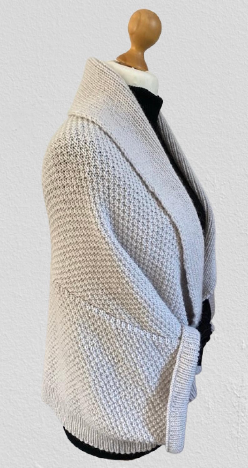 Op de afbeelding: Een beige gebreid vest met een brede sjaalkraag en een gestructureerd breipatroon. Het kledingstuk heeft lange mouwen met geribbelde boorden en een zoom die op de taille valt, gedragen over een zwarte top met hoge hals op een etalagepop.