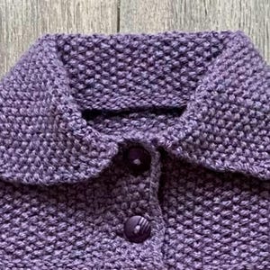 May include: Close-up of a knitted purple sweater with a rounded collar and two matching buttons. The textured knit fabric is visible, showcasing the craftsmanship of the garment. The sweater is likely handmade.