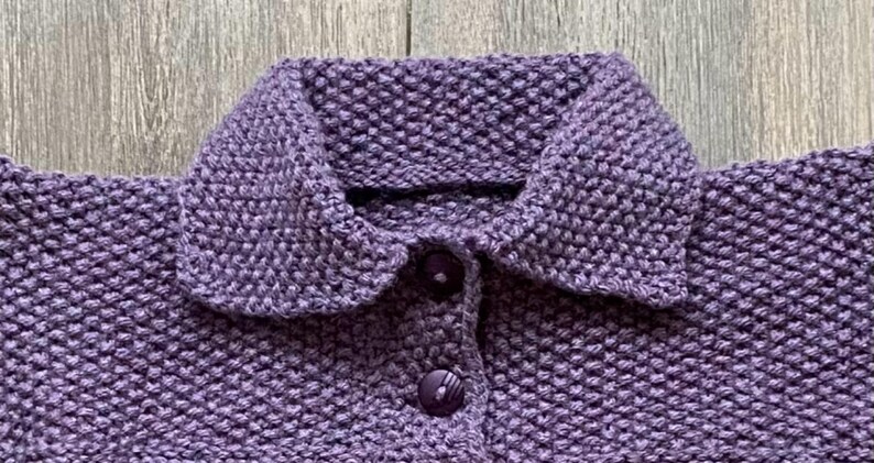 May include: Close-up of a knitted purple sweater with a rounded collar and two matching buttons. The textured knit fabric is visible, showcasing the craftsmanship of the garment. The sweater is likely handmade.