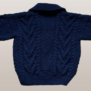 May include: A dark navy blue long-sleeve sweater featuring a prominent cable knit pattern throughout the body and sleeves. The garment includes a wide fold-over collar and ribbed cuffs at the wrists and hem, showcasing a thick, textured knit construction.