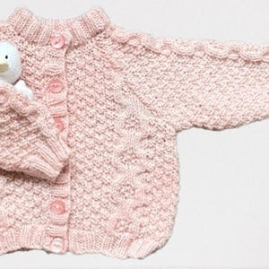 Op de afbeelding: Een lichtroze gebreid babyvestje met een knoopsluiting aan de voorkant en een klein wit stoffen eendje in de zak. Het vestje heeft een textuur gebreid patroon en is gemaakt van zacht garen.