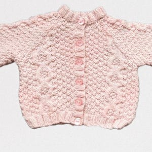 Op de afbeelding: Een lichtroze gebreid babyvestje met een ronde kraag en lange mouwen. De trui heeft een textuurpatroon en wordt gesloten met vijf roze knopen. De mouwen hebben een kabelgebreid ontwerp.