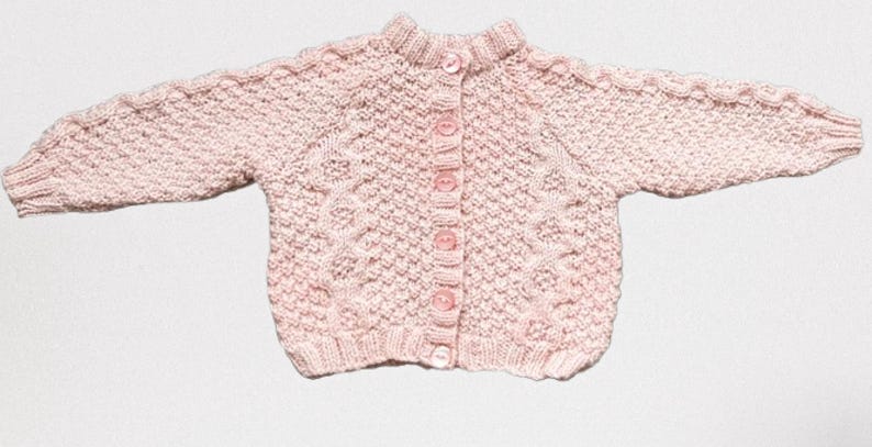 Op de afbeelding: Een lichtroze gebreid babyvestje met een ronde kraag en lange mouwen. De trui heeft een textuurpatroon en wordt gesloten met vijf roze knopen. De mouwen hebben een kabelgebreid ontwerp.