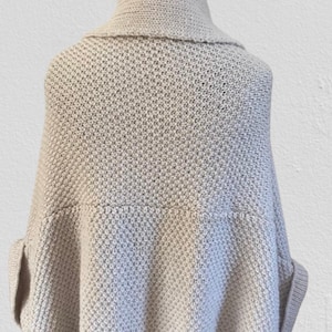 Op de afbeelding: Een lichtbeige gebreide poncho met een brede omgeslagen kraag en een gestructureerd breipatroon. Het kledingstuk heeft een losvallend, gedrapeerd silhouet met mouwboorden en een geribbelde zoom voor een gestructureerde en ontspannen uitstraling.