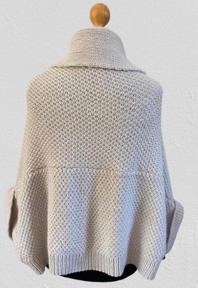 Op de afbeelding: Een lichtbeige gebreide poncho met een brede omgeslagen kraag en een gestructureerd breipatroon. Het kledingstuk heeft een losvallend, gedrapeerd silhouet met mouwboorden en een geribbelde zoom voor een gestructureerde en ontspannen uitstraling.