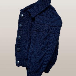 May include: A dark navy blue long-sleeve cardigan featuring a thick cable knit pattern throughout the body and sleeves. The garment has a folded collar, a ribbed hem, and a vertical row of five matching dark navy blue circular buttons down the front.