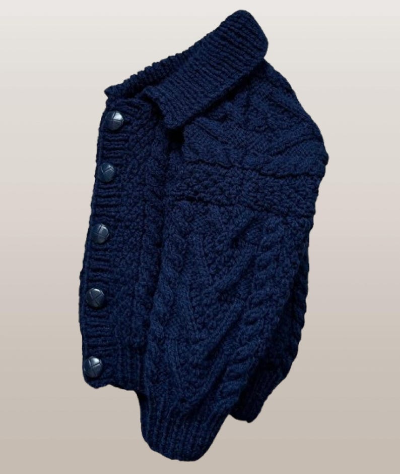 May include: A dark navy blue long-sleeve cardigan featuring a thick cable knit pattern throughout the body and sleeves. The garment has a folded collar, a ribbed hem, and a vertical row of five matching dark navy blue circular buttons down the front.