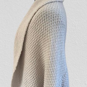 Op de afbeelding: Een lichtgrijs gebreid vest met een textuur in wafelsteek, een brede omgeslagen kraag en mouwen met boorden. Het kledingstuk wordt getoond op een etalagepop en heeft een gedrapeerd, open silhouet met geribbelde details langs de onderzoom.