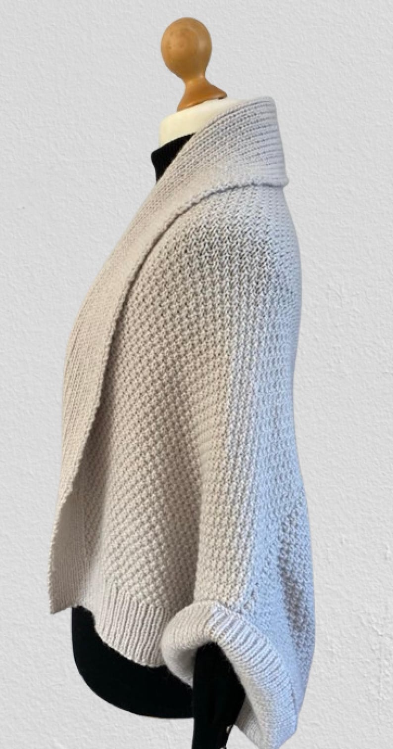 Op de afbeelding: Een lichtgrijs gebreid vest met een textuur in wafelsteek, een brede omgeslagen kraag en mouwen met boorden. Het kledingstuk wordt getoond op een etalagepop en heeft een gedrapeerd, open silhouet met geribbelde details langs de onderzoom.