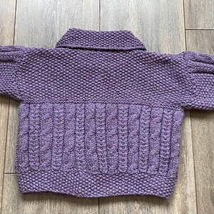 May include: A lavender-colored, hand-knitted sweater with a collared neckline and cable knit detailing on the lower front. The sleeves are a puffy style. The sweater is lying flat on a wooden surface.