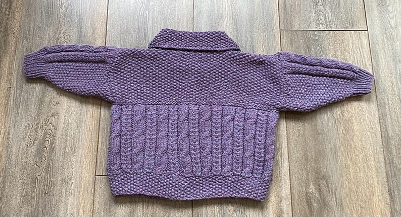 May include: A lavender-colored, hand-knitted sweater with a collared neckline and cable knit detailing on the lower front. The sleeves are a puffy style. The sweater is lying flat on a wooden surface.