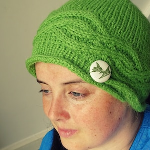 May include: A green knitted hat with a unique cable knit pattern and a button with a bird design. The hat has a wide brim and is worn by a person with dark hair.