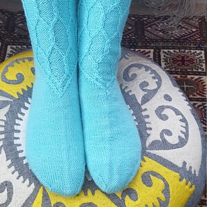 Op de afbeelding: Een paar turquoise blauwe gebreide sokken met een ruitpatroon. De sokken worden gedragen op de benen van een persoon en rusten op een grijs en geel gepatroneerd kussen.