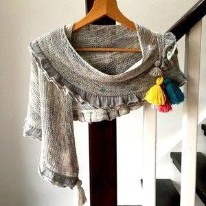 May include: A gray knitted shawl with a ruffled edge and colorful tassels. The shawl is hanging on a wooden hanger.