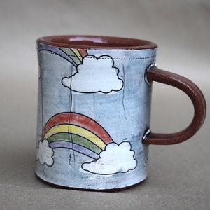 May include: A ceramic mug with a light blue background and a rainbow and cloud design. The mug has a brown handle.