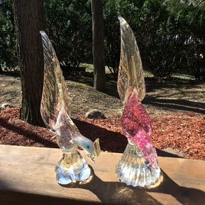 May include: Two glass bird sculptures with iridescent wings. The bird on the left is clear with a light pink hue and a green eye. The bird on the right is pink with a clear base and a green eye.