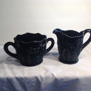 Puede incluir: Un juego de azucarero y cremera de vidrio negro. El azucarero tiene dos asas y un borde festoneado. La cremera tiene un asa y un pico. Ambas piezas tienen un patrón detallado en relieve. El juego se exhibe sobre una superficie blanca.