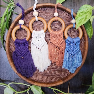 May include: Four macrame car charms in purple, white, copper, and blue, each with a wooden ring and bead. The charms are displayed on a woven tray, showcasing the handmade details and color variations.