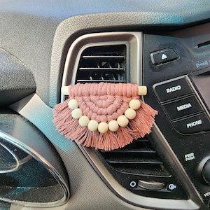 May include: A dusty rose macrame car air freshener with a half-circle design. The air freshener features a row of light wooden beads and a fringe. It is attached to a car vent.