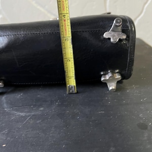 May include: A black leather handbag base featuring four silver-toned metal feet. A yellow measuring tape is positioned vertically against the side, indicating a height of approximately 4 inches for the base of the bag.