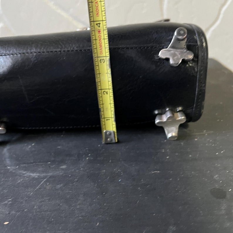 May include: A black leather handbag base featuring four silver-toned metal feet. A yellow measuring tape is positioned vertically against the side, indicating a height of approximately 4 inches for the base of the bag.