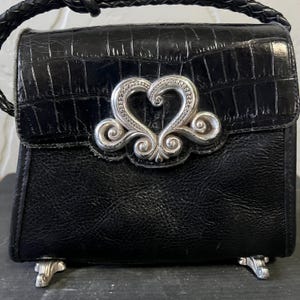 May include: A black handbag with a textured crocodile-embossed flap and a smooth leather body. The bag features a prominent silver-toned metal heart-shaped ornament on the front, a braided leather handle, and four silver-toned metal feet at the base.