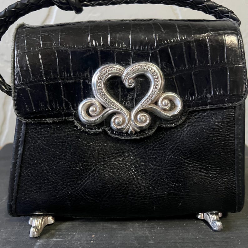May include: A black handbag with a textured crocodile-embossed flap and a smooth leather body. The bag features a prominent silver-toned metal heart-shaped ornament on the front, a braided leather handle, and four silver-toned metal feet at the base.
