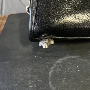 May include: A black textured leather handbag corner featuring a silver-toned metal protective foot at the base. The material has a visible grain pattern, and a metallic ring attachment is partially visible at the top edge of the bag structure.