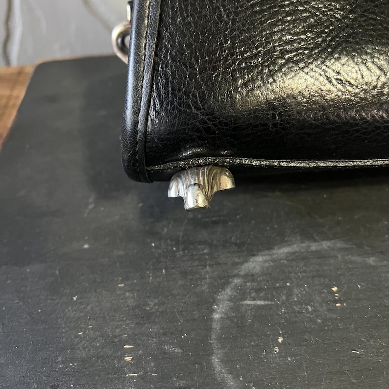 May include: A black textured leather handbag corner featuring a silver-toned metal protective foot at the base. The material has a visible grain pattern, and a metallic ring attachment is partially visible at the top edge of the bag structure.