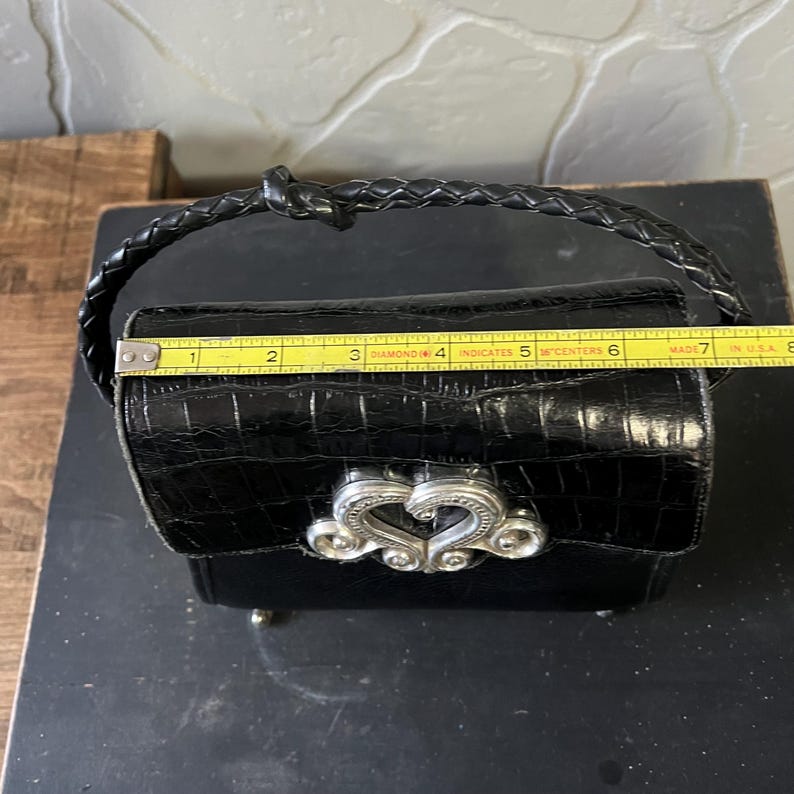 May include: A black handbag with a crocodile-embossed texture, a braided handle, and a silver-toned heart-shaped metal clasp. A yellow measuring tape is placed across the top, showing a width of approximately 7 inches for the bag's main body.