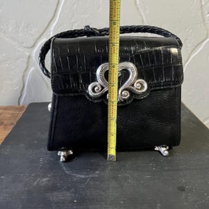 May include: A black handbag with a crocodile-embossed flap and a braided handle. A silver-toned ornate metal clasp is centered on the front. A yellow measuring tape placed vertically shows the bag is approximately 8 inches tall from base to top.