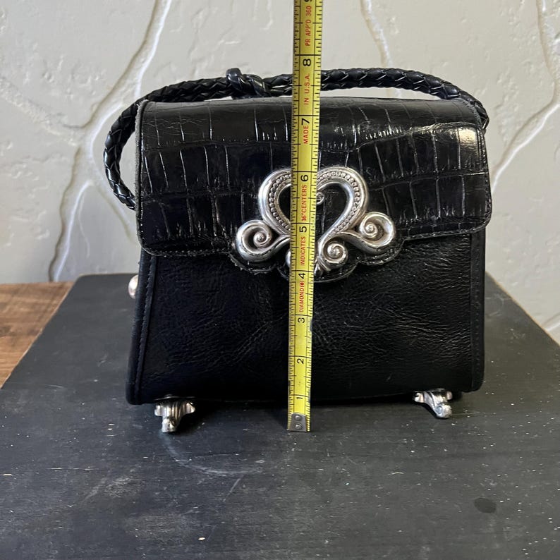 May include: A black handbag with a crocodile-embossed flap and a braided handle. A silver-toned ornate metal clasp is centered on the front. A yellow measuring tape placed vertically shows the bag is approximately 8 inches tall from base to top.