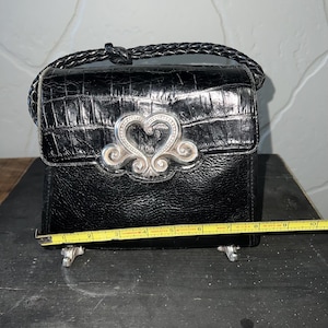 May include: A black handbag with a textured finish and a braided handle. A silver-toned heart-shaped ornament is centered on the front flap. A yellow measuring tape is positioned across the base, showing a width of approximately 9.5 inches.
