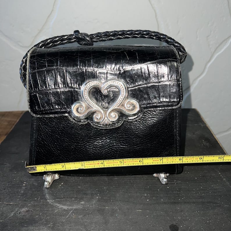 May include: A black handbag with a textured finish and a braided handle. A silver-toned heart-shaped ornament is centered on the front flap. A yellow measuring tape is positioned across the base, showing a width of approximately 9.5 inches.