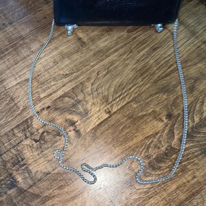 May include: A black textured leather handbag featuring a long silver-toned metal chain shoulder strap and two small silver-toned metal feet attached to the bottom edge of the bag, providing a stable base for the accessory when placed upright.