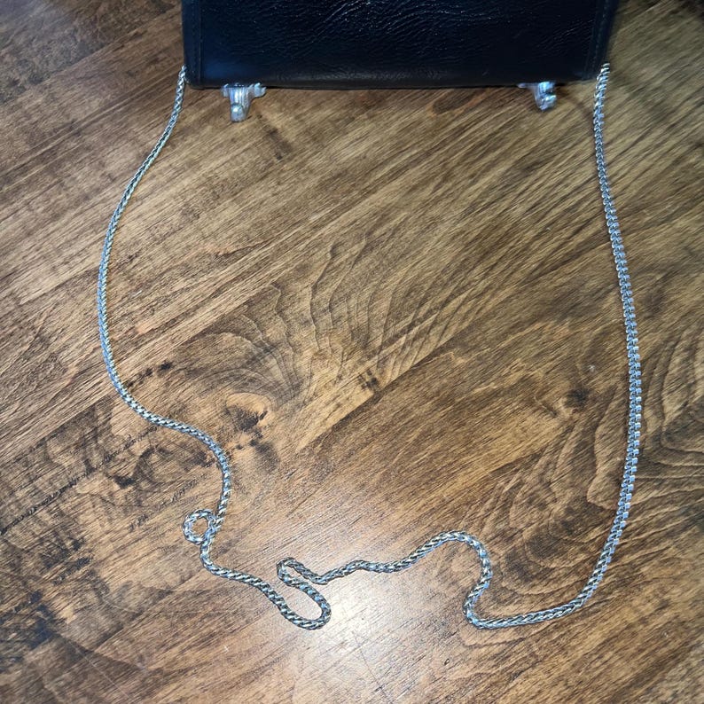 May include: A black textured leather handbag featuring a long silver-toned metal chain shoulder strap and two small silver-toned metal feet attached to the bottom edge of the bag, providing a stable base for the accessory when placed upright.