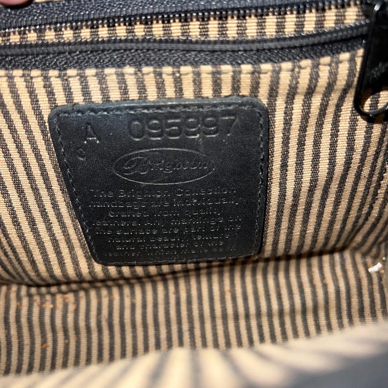 May include: The interior of a handbag featuring a black leather patch embossed with the Brighton logo and the serial number 095997. The lining consists of a beige and black vertical striped fabric. The text on the patch describes the collection.