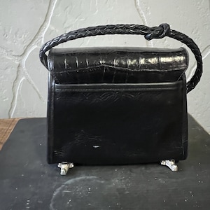 May include: A black handbag with a crocodile-embossed flap and a braided handle. The bag features a smooth black body, a horizontal pocket on the front, and four silver-toned metal feet at the base for stability when placed on a flat surface.