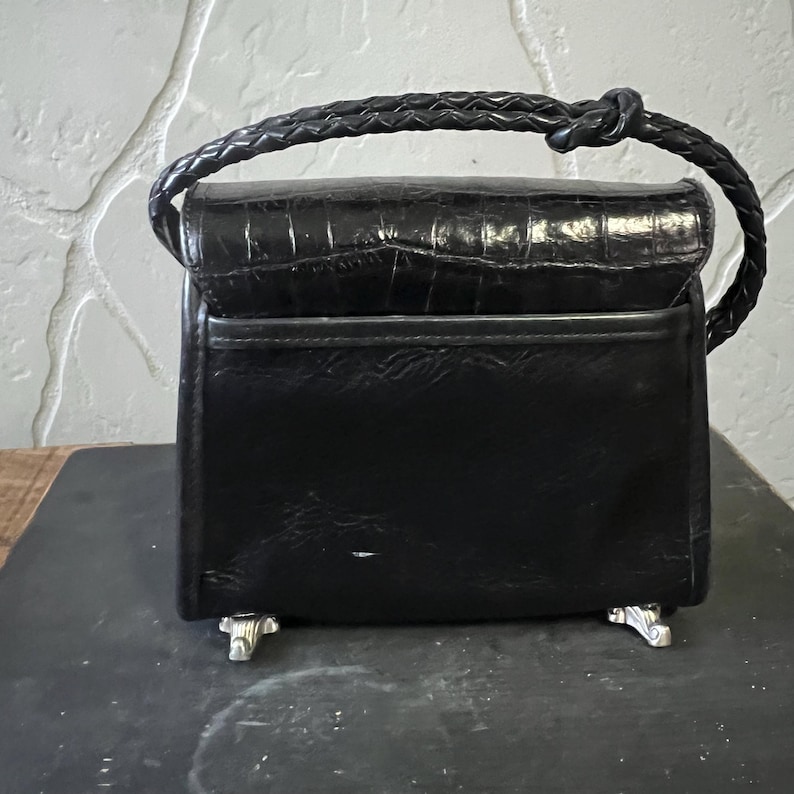 May include: A black handbag with a crocodile-embossed flap and a braided handle. The bag features a smooth black body, a horizontal pocket on the front, and four silver-toned metal feet at the base for stability when placed on a flat surface.